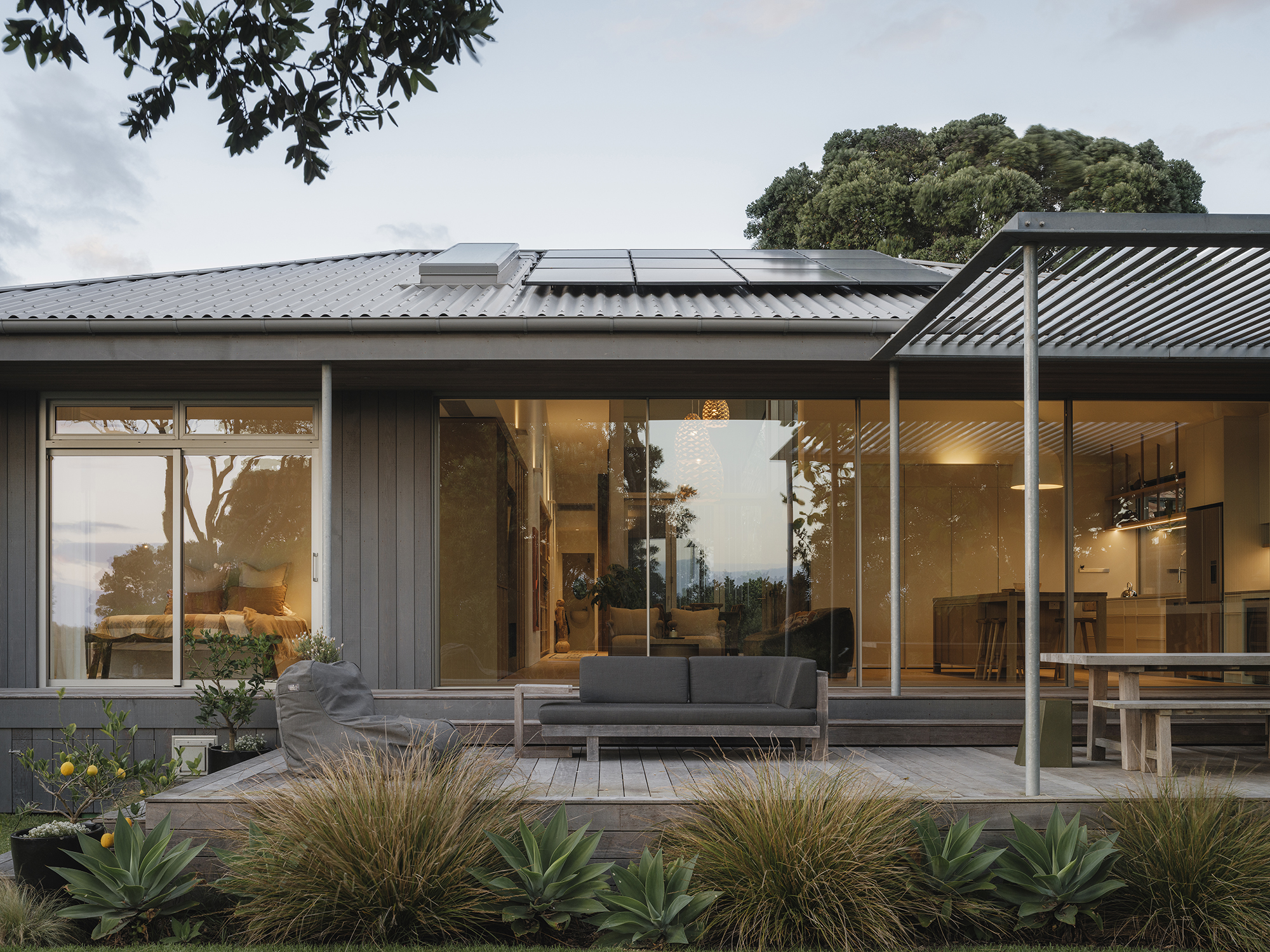 Treefold residence at dusk — gabled timber pavilion opening onto a north-facing court, a Bay of Plenty coastal context